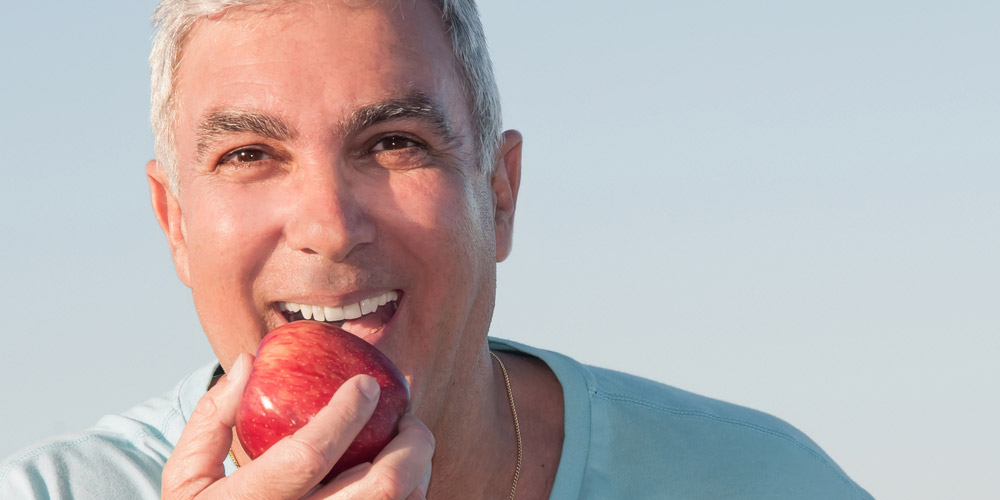 A man is eating an apple in front of a blue sky.